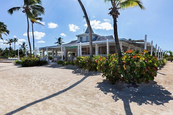 A beach club with a covered seating area, palm trees, and a sandy beach.