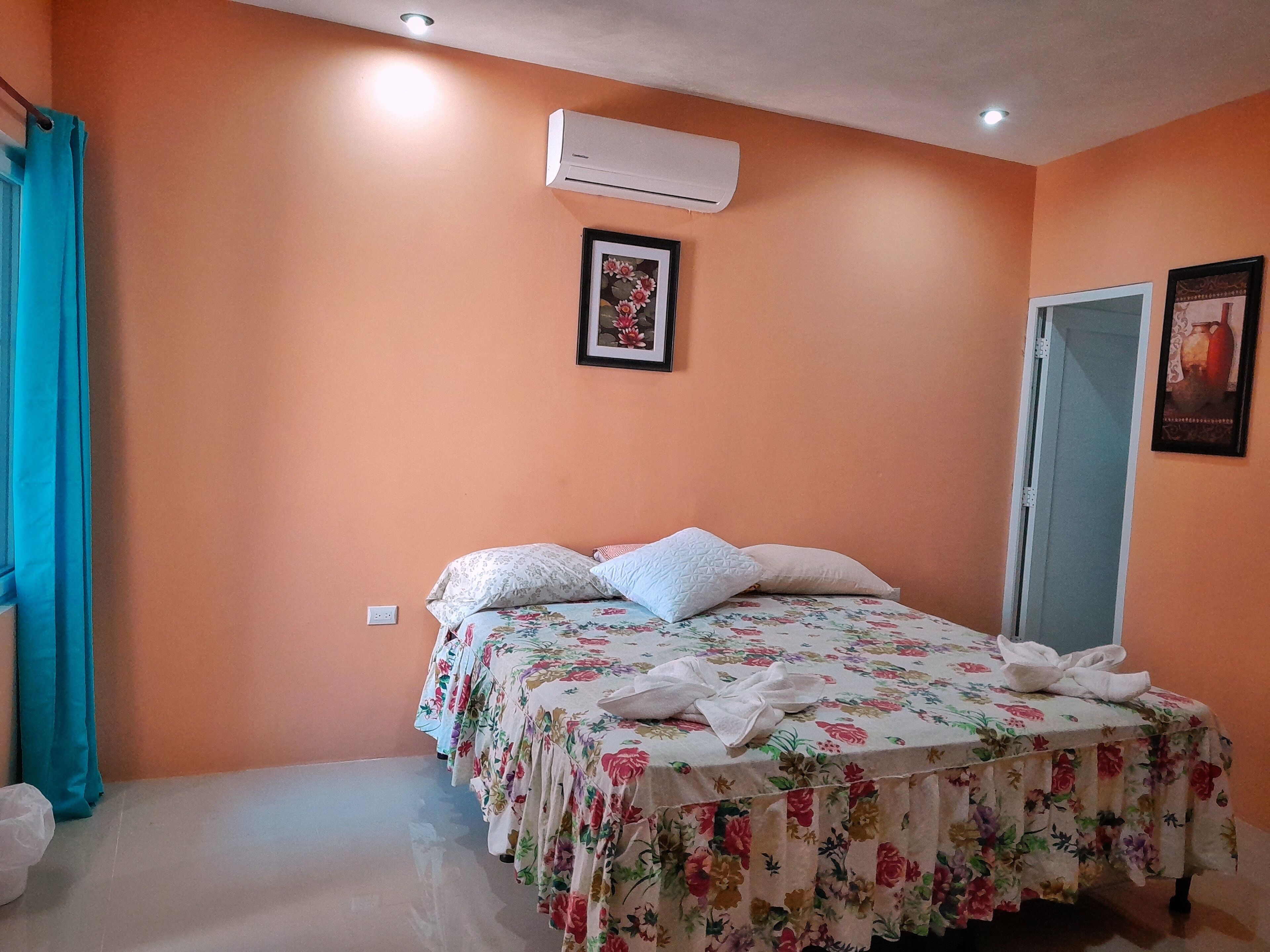 A bedroom with a floral bedspread, a single bed with pillows, an air conditioning unit, and a framed picture on the wall.