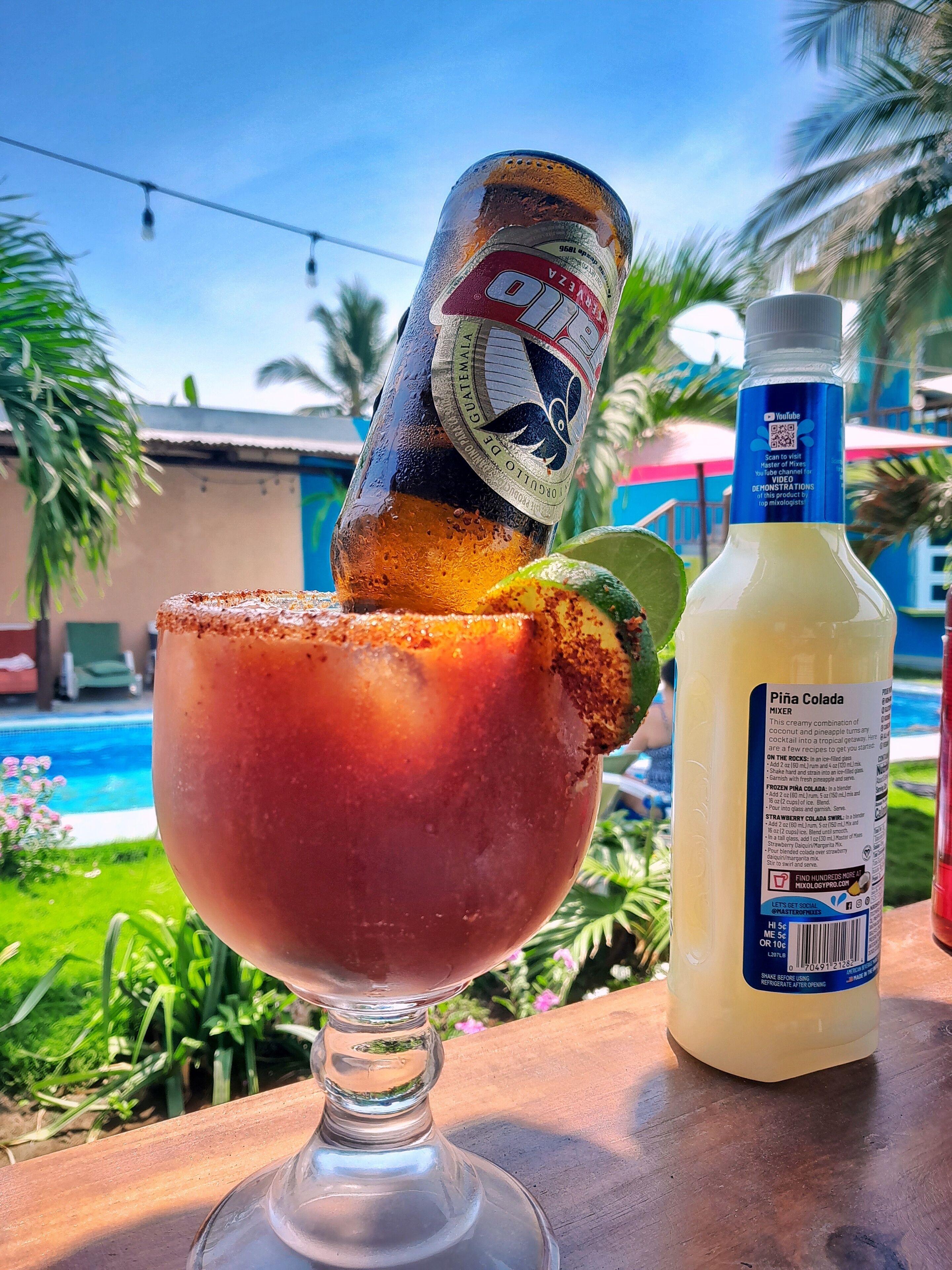 A tropical drink in a glass with a lime wedge, a bottle of Piña Colada mixer, and a bottle of clear liquid, possibly a mixer or soda, on a wooden table with a pool and palm trees in the background.
