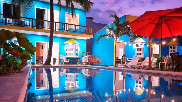 A swimming pool with lounge chairs and umbrellas, surrounded by buildings with balconies and large windows. - Hotel Evenezer El Paredon (Sipacate)