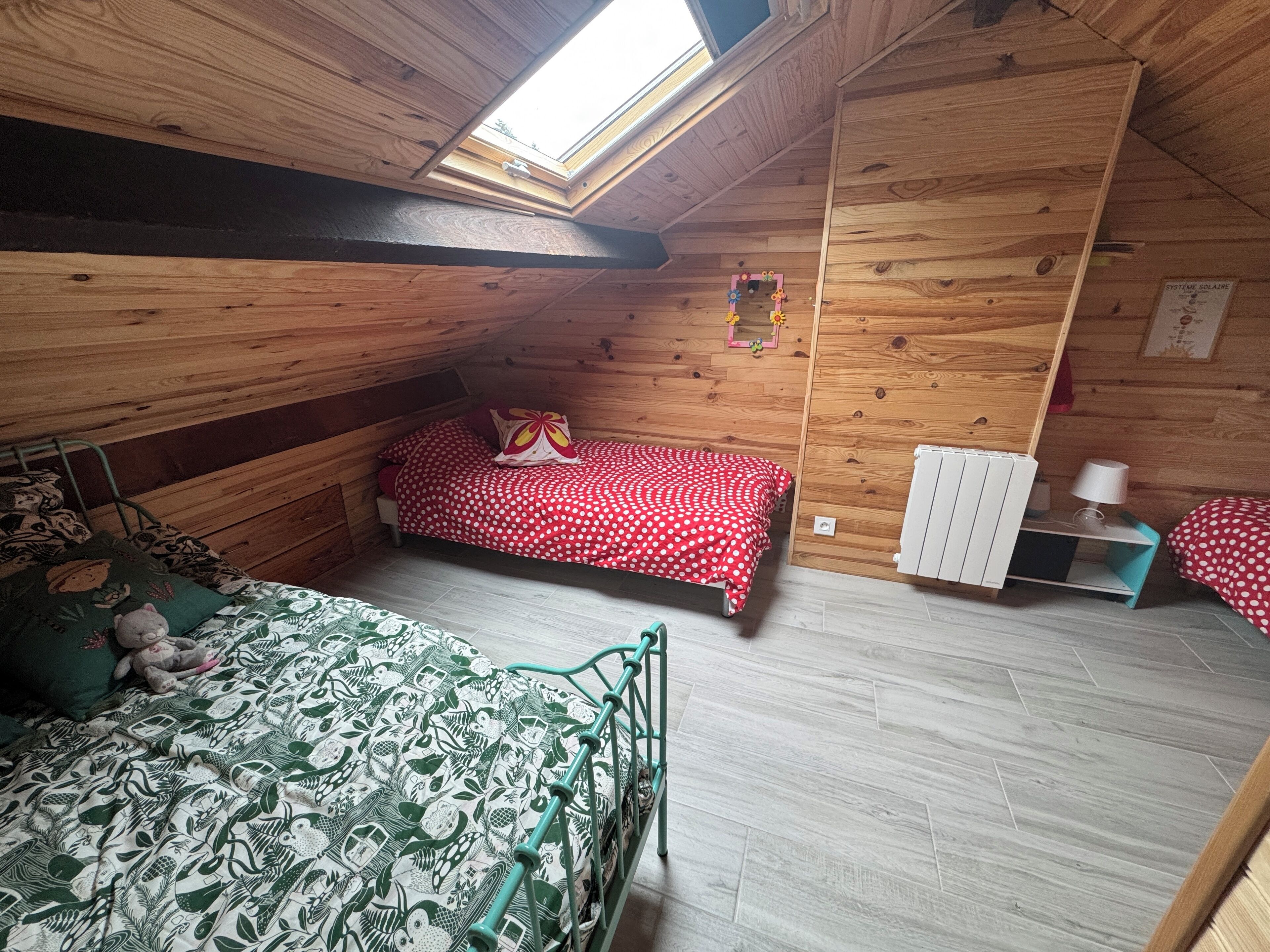 Une chambre mansardée chaleureuse, avec un plafond en pente, un puits de lumière, un lit recouvert d'une couvre-lit à motifs, un plus petit lit avec une couvre-lit à pois, un radiateur et une table de chevet avec une lampe.