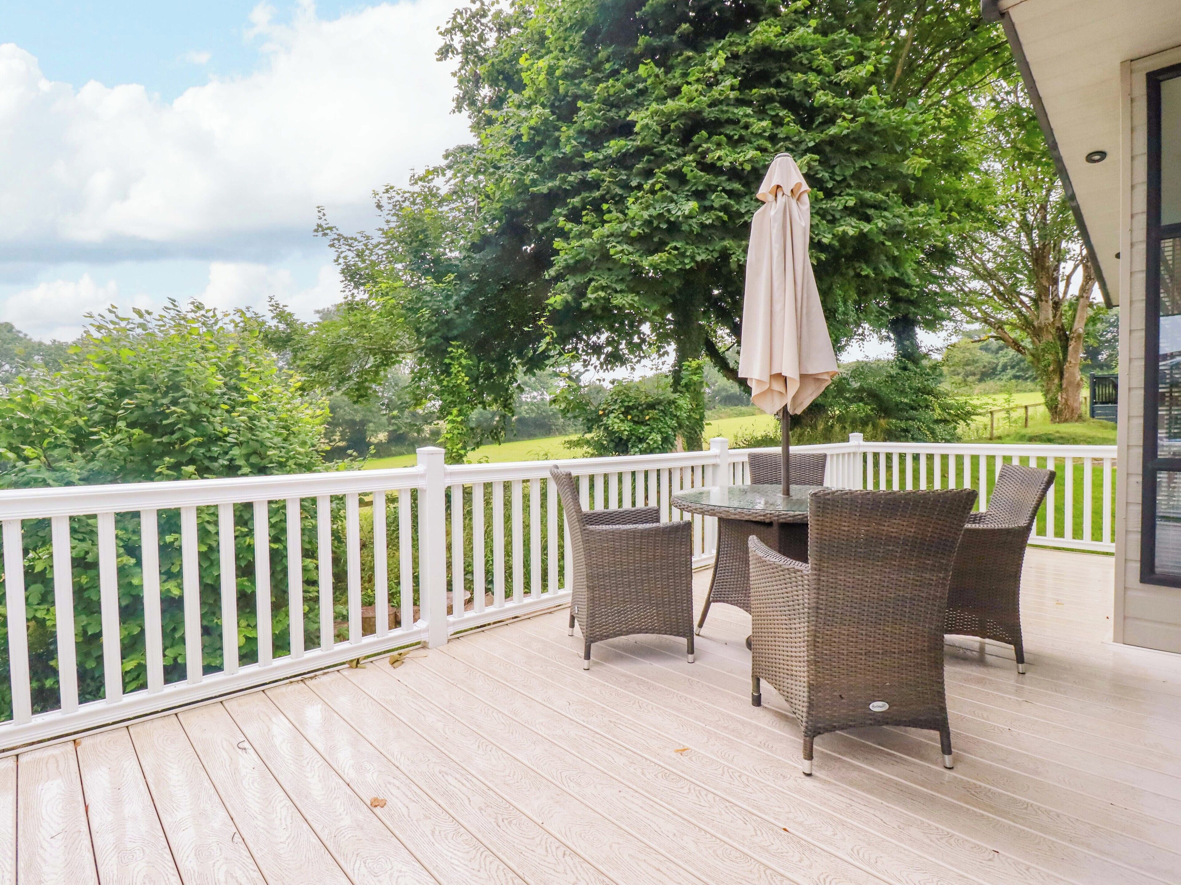 A covered outdoor deck with wicker furniture and a table, surrounded by a white railing and a large tree.