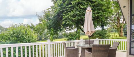 A covered outdoor deck with wicker furniture and a table, surrounded by a white railing and a large tree.