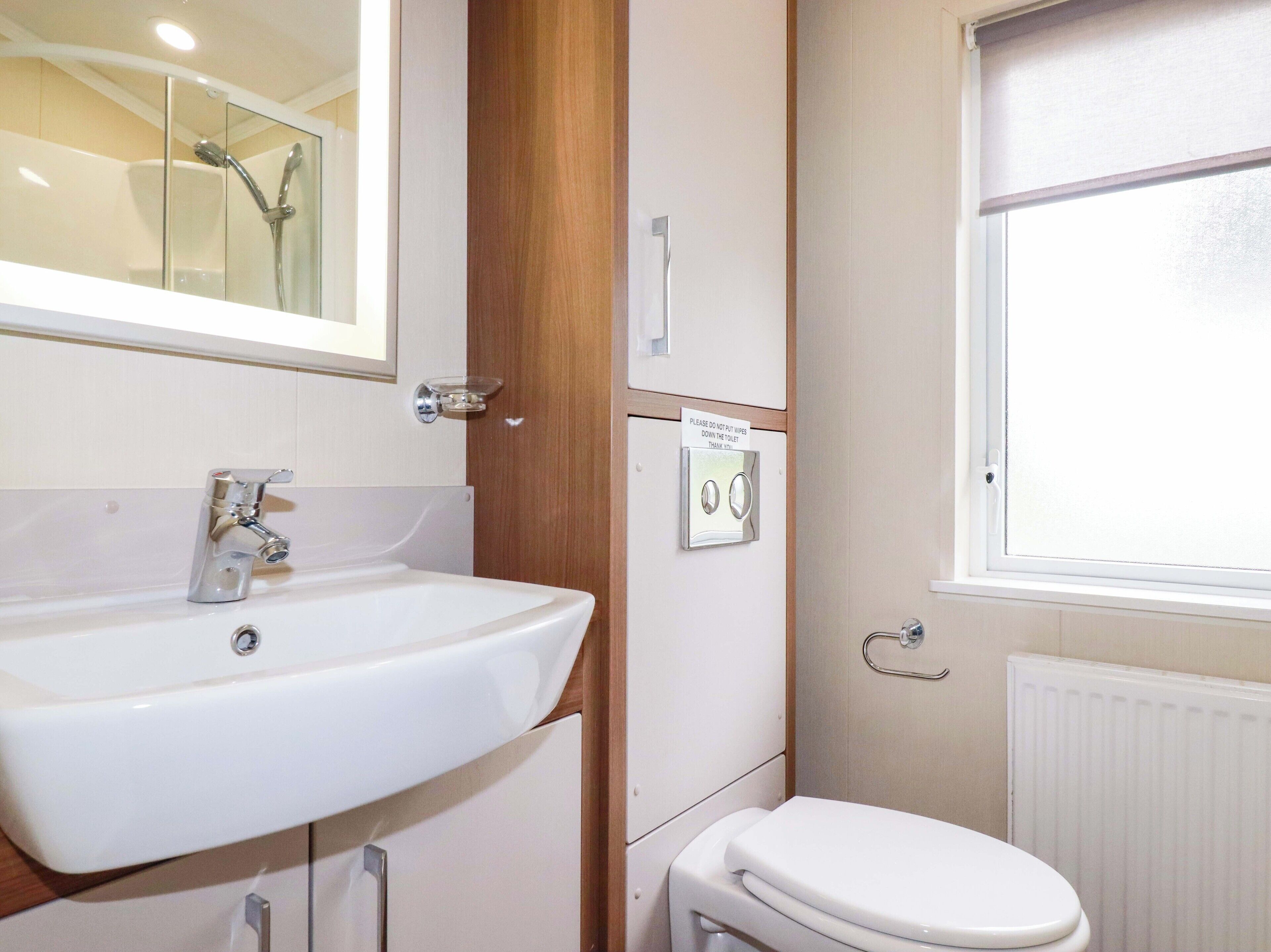 Een badkamer met een witte wasbak, een toilet en een douche. Op de muur hangt een bordje met de tekst 'Wipes niet doorspoelen'.