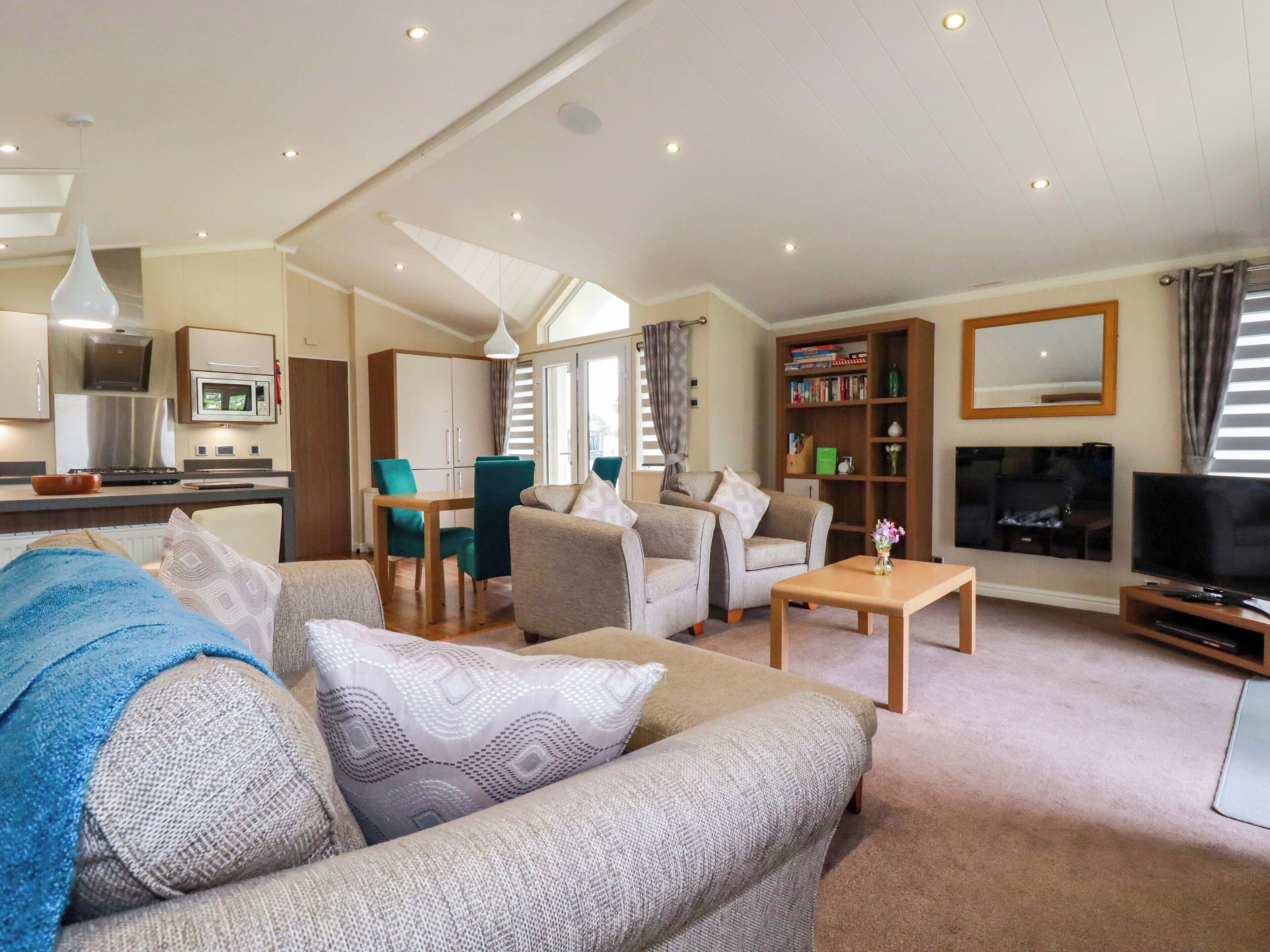 A modern living room with a sofa, chairs, a coffee table, a bookshelf, a TV, and a kitchen area with a microwave and cabinets.