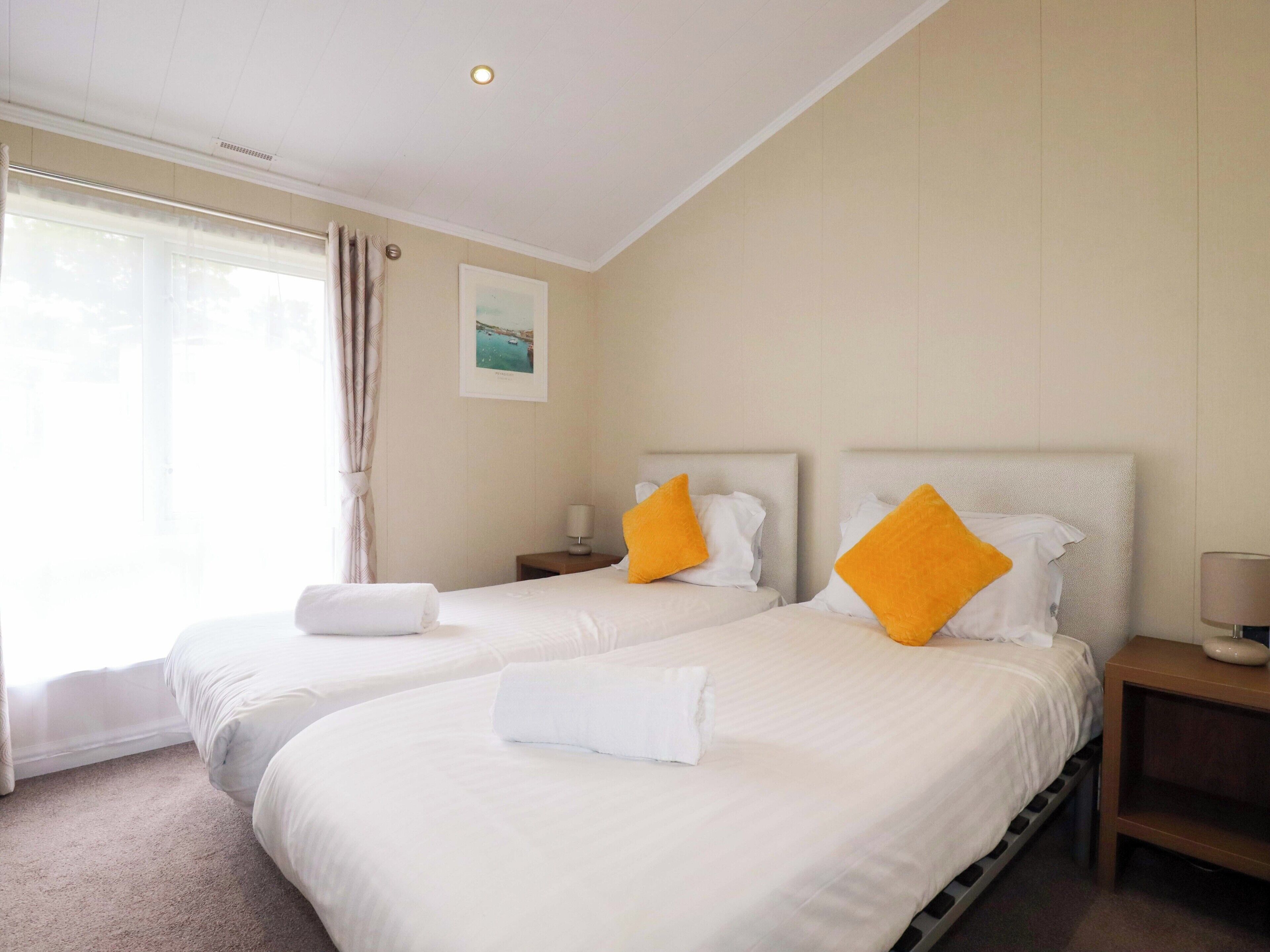Une chambre d’hôtel avec deux lits, chacun agrémenté de draps blancs et d’oreillers orange, une petite table de chevet avec une lampe, et une fenêtre avec des rideaux légers.