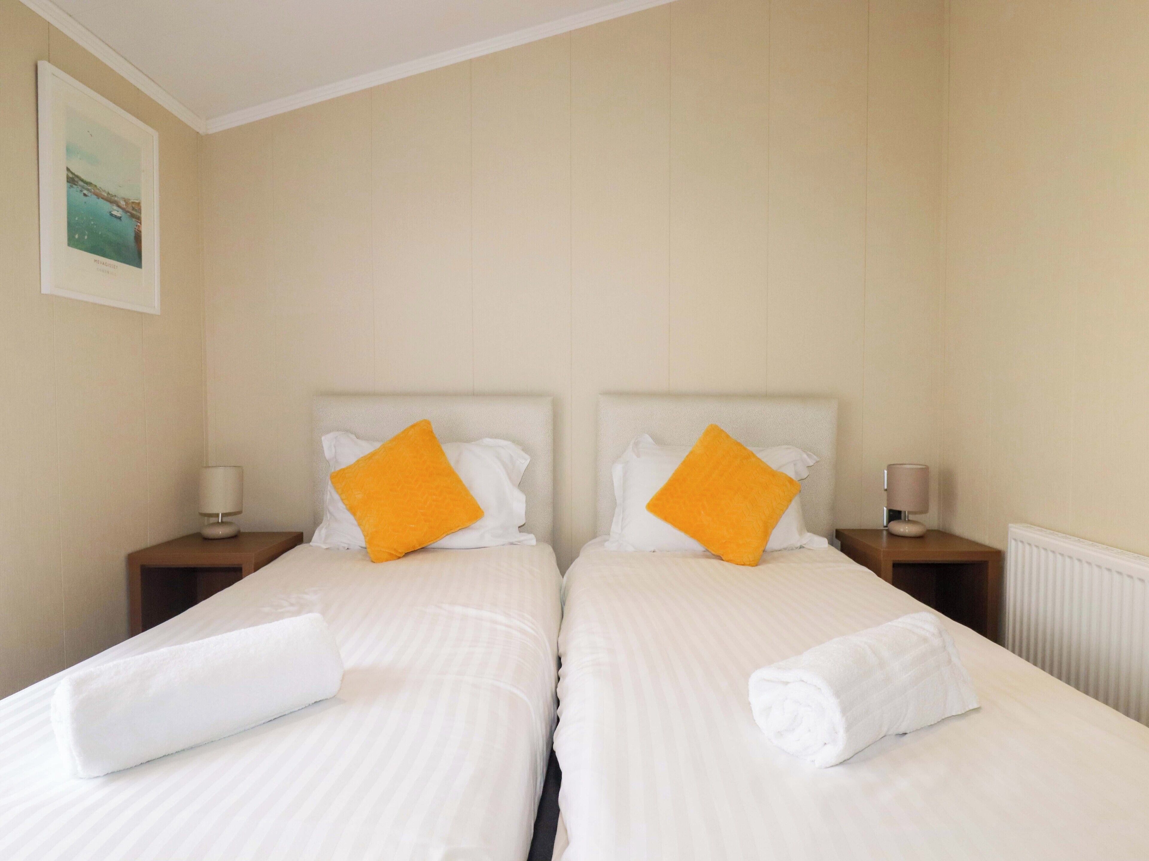A double bed with white linens and two orange pillows, flanked by nightstands with lamps, in a room with beige walls and a framed picture on the wall.