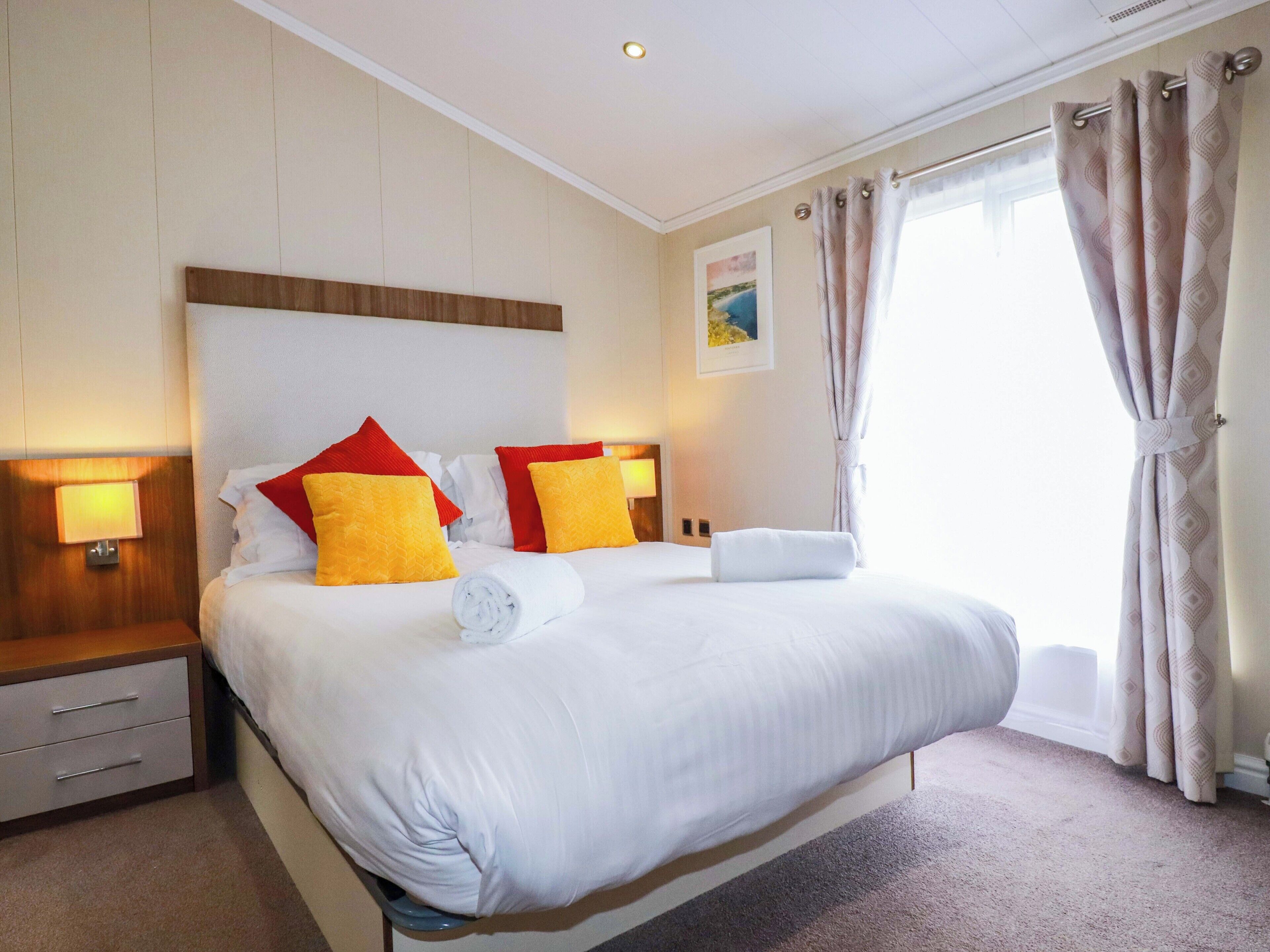 A hotel room with a large bed, two bedside tables, a headboard, a window with curtains, and a wall-mounted lamp.