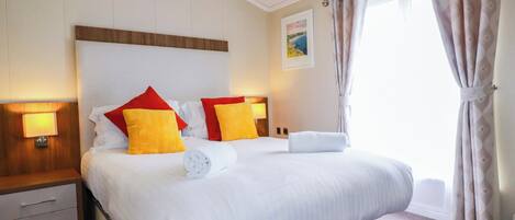 A hotel room with a large bed, two bedside tables, a headboard, a window with curtains, and a wall-mounted lamp.