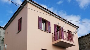 A two-story building with a balcony, a small garden, and a stone wall. - ΜΟΝΟΚΑΤΟΙΚΙΑ ΣΕ ΠΑΡΑΔΟΣΙΑΚΟ ΟΙΚΙΣΜΟ (ΜΥΡΙΝΑ ΛΗΜΝΟΥ)