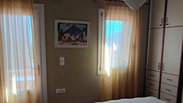 A bedroom with a bed, curtains, a painting, and a wardrobe. - ΜΟΝΟΚΑΤΟΙΚΙΑ ΣΕ ΠΑΡΑΔΟΣΙΑΚΟ ΟΙΚΙΣΜΟ (ΜΥΡΙΝΑ ΛΗΜΝΟΥ)
