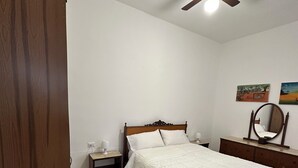 Een slaapkamer met een houten bed, een nachtkastje, een dressoir, een plafondventilator en twee aan de muur bevestigde schilderijen.