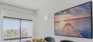 Un comedor con una mesa de madera preparada para el desayuno, un cuenco de fruta y un plato de cruasanes. Un amplio ventanal ofrece vistas a un cuerpo de agua y edificios en la distancia.