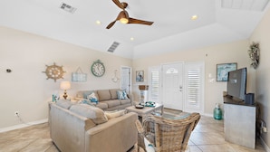 A spacious living room with a beige sofa, a wicker chair, a ceiling fan, and a television on a stand. - Purple Pelican Paradise by Ryson Vacation Rentals (Galveston)