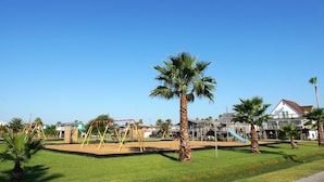 A playground with swings and a slide, surrounded by palm trees and a building with a balcony. - Parijatha by Ryson Vacation Rentals (Galveston)