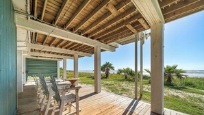 A wooden deck with chairs and a table, overlooking a beach with palm trees and the ocean. - Parijatha by Ryson Vacation Rentals (Galveston)
