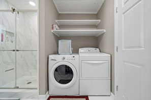 A compact laundry room with a washing machine, dryer, and a glass-enclosed shower. - Parijatha by Ryson Vacation Rentals (Galveston)