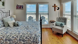 A bedroom with a large bed, a chair, a cross on the wall, and a window with blinds. - Casablanca by Ryson Vacation Rentals (Galveston)