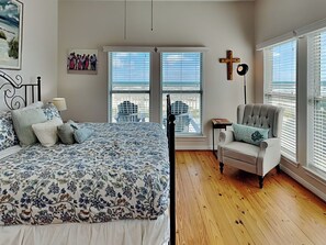 Une chambre à coucher avec un grand lit, une chaise, un crucifix au mur et une fenêtre avec des stores.
