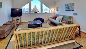 A spacious living room with a high ceiling, a large window with a sailboat view, a flat-screen TV, a wooden sofa, and a small table. - Casablanca by Ryson Vacation Rentals (Galveston)