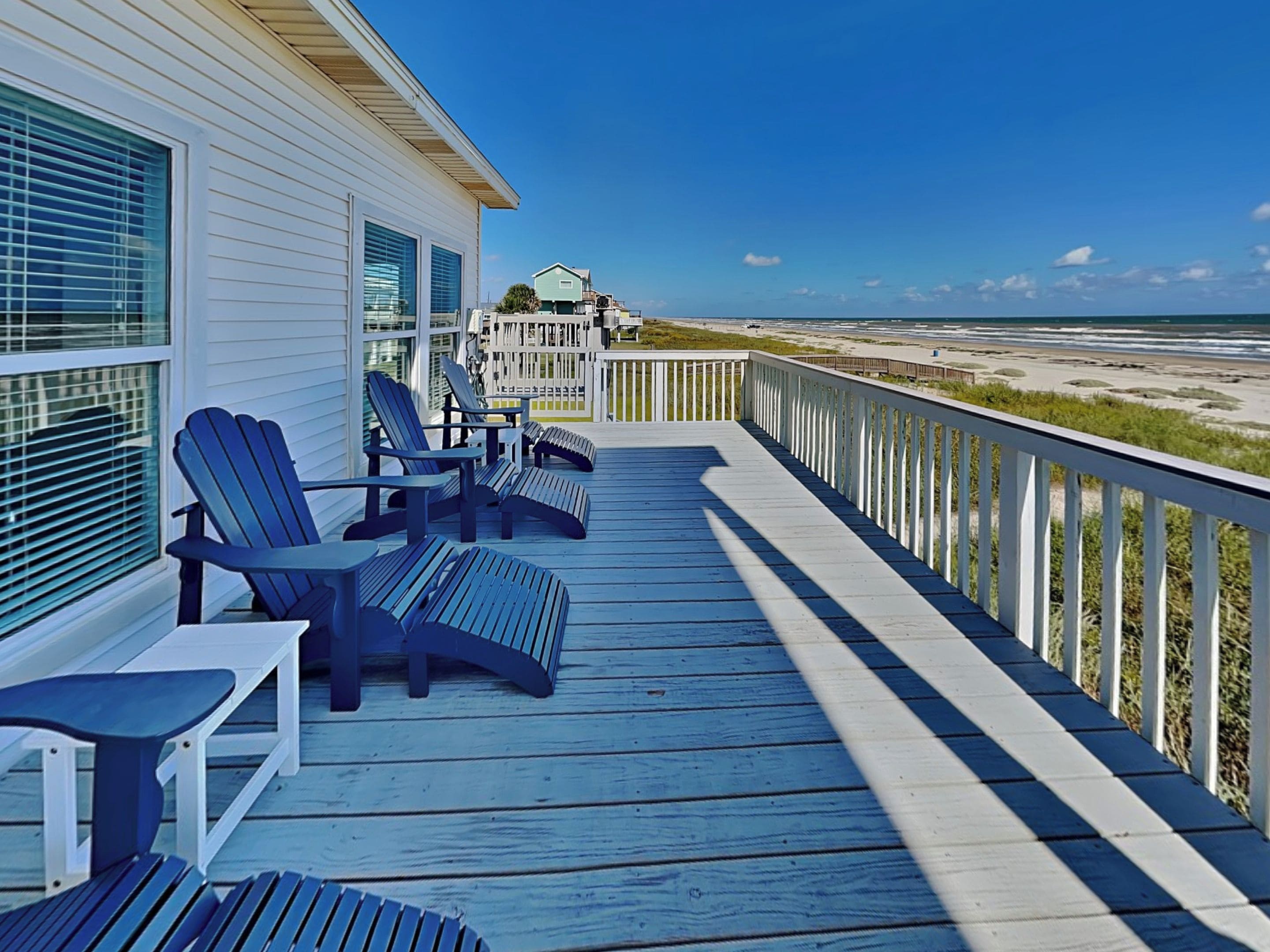 Een houten terras met Adirondack stoelen, een witte balustrade en uitzicht op het strand.