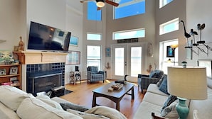 A spacious living room with a fireplace, comfortable seating, a television, and large windows offering views. - Casablanca by Ryson Vacation Rentals (Galveston)