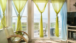 A room with a view of the beach, a wicker chair, a television, and a window with curtains. - Beachfront Melody by Ryson Vacation Rentals (Galveston)