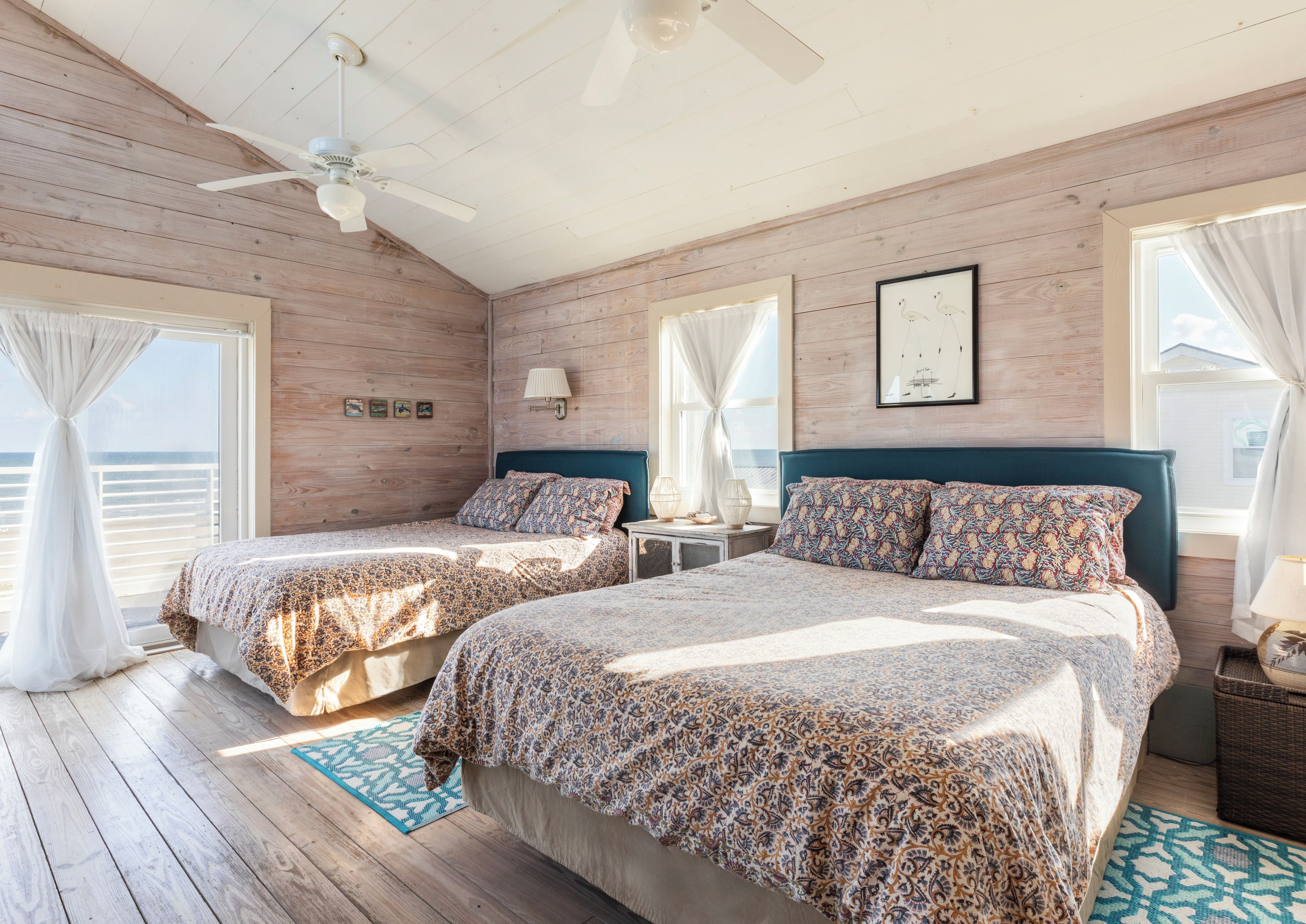 A bedroom with two beds, wooden walls, a ceiling fan, and a view of the sea.