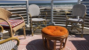 A deck with wicker chairs and a round ottoman, overlooking a beach. - Beachfront Melody by Ryson Vacation Rentals (Galveston)