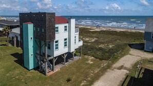 A two-story building with a unique architectural design, featuring a combination of white and dark-colored walls, and a red roof. The building is situated on a grassy area with a view of the ocean and beach in the background. - Beachfront Melody by Ryson Vacation Rentals (Galveston)