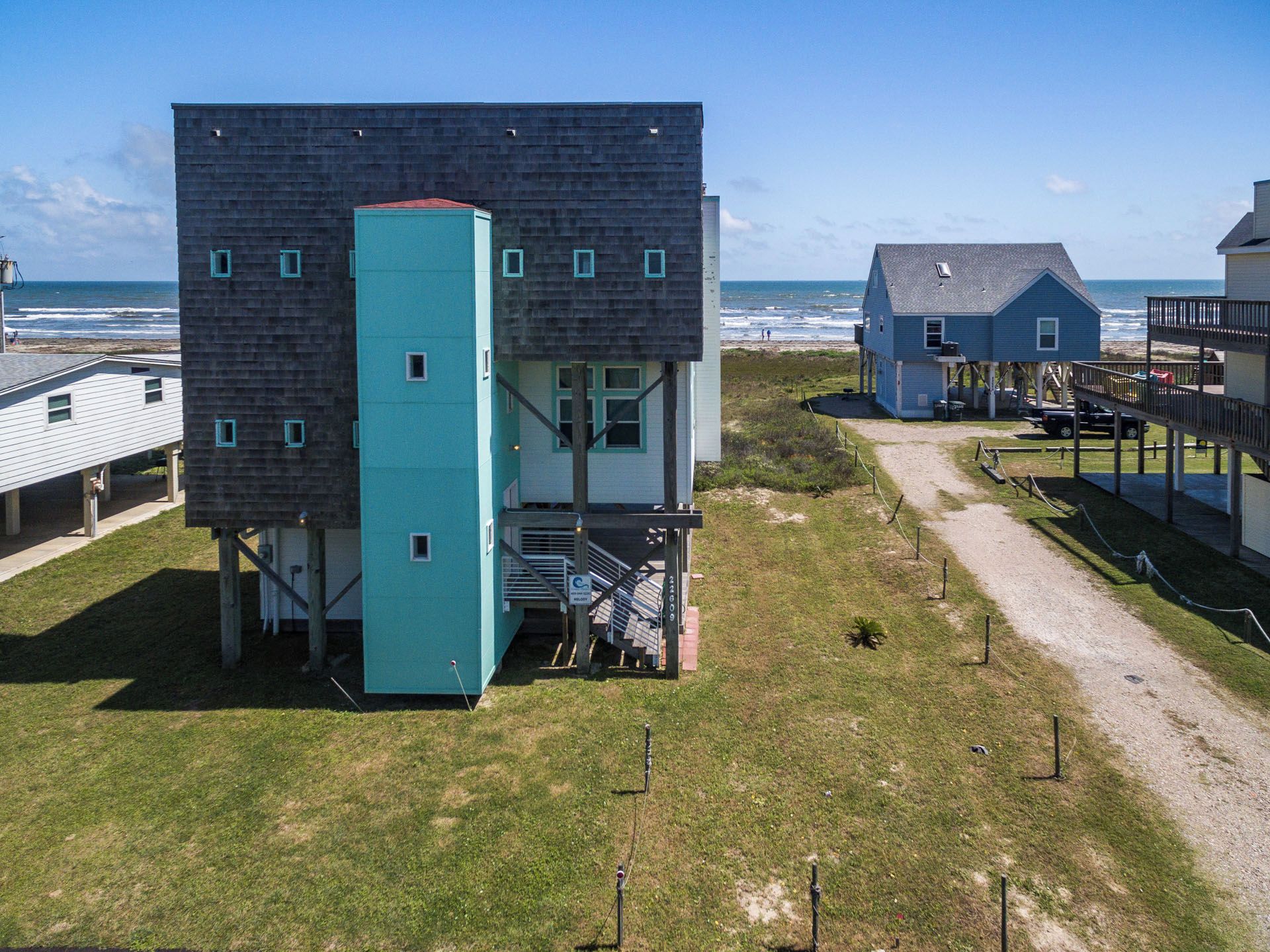A coastal building with a staircase leading to a deck, surrounded by a grassy area and a beach in the background.