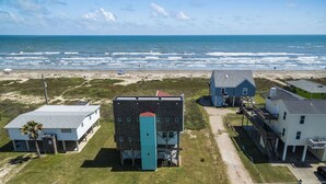 A coastal building complex with a beach view, including a prominent blue structure with a red roof, adjacent white and green buildings, and a clear path leading to the beach. - Beachfront Melody by Ryson Vacation Rentals (Galveston)