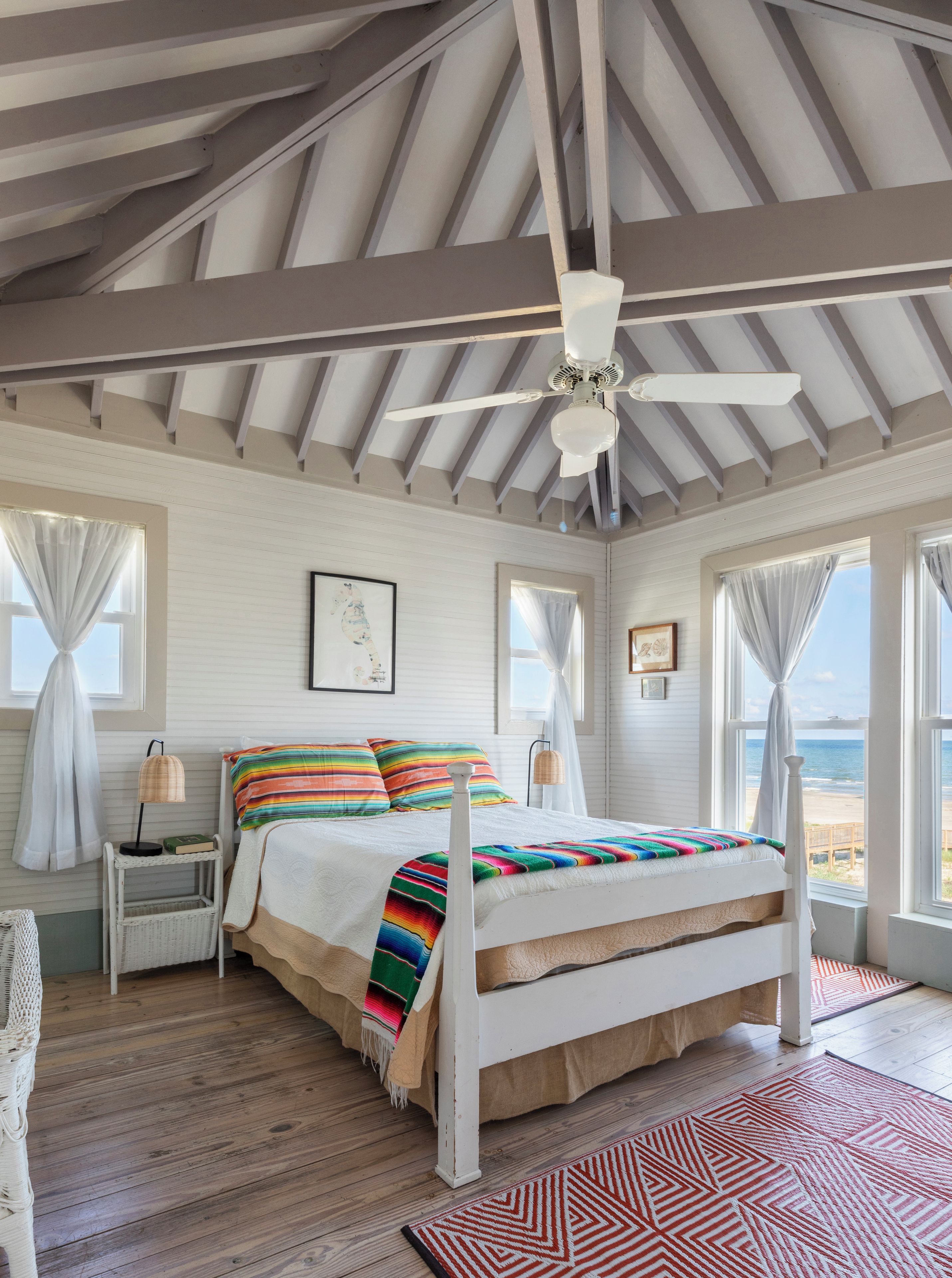 A bedroom with a bed, a ceiling fan, a nightstand, a lamp, and a view of the beach.
