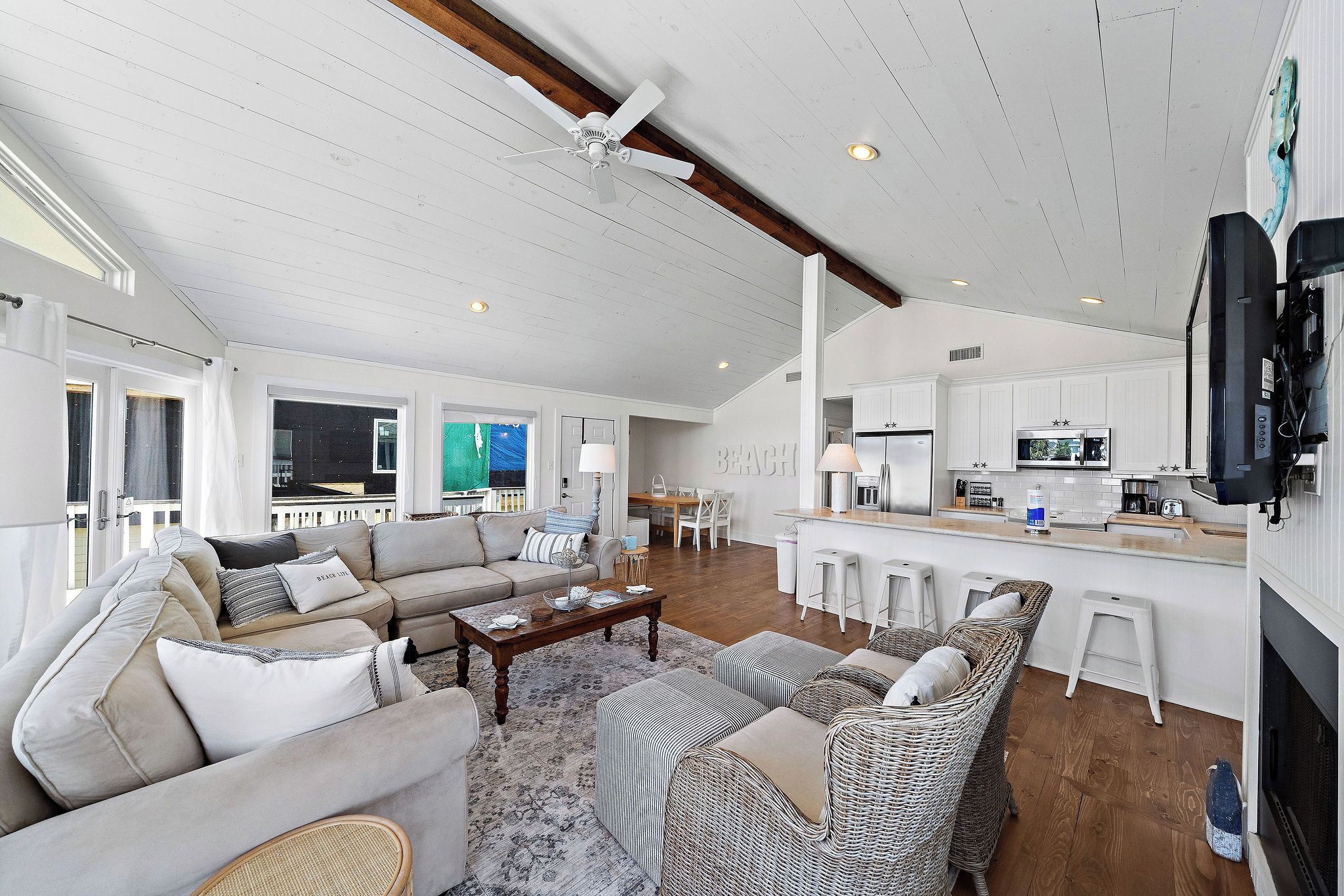 A spacious living area with a sofa set, a coffee table, and a wicker chair. A kitchen area with a bar and stools is visible in the background.