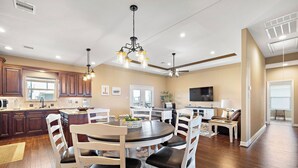 A modern kitchen with dark wooden cabinets, a dining area with white chairs, a ceiling fan, and a large window. - Copper Top by Ryson Vacation Rentals (Galveston)
