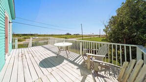 A wooden deck with a table and chairs, overlooking a grassy area with a road and distant buildings. - Colva Tides by Ryson Vacation Rentals (Galveston)