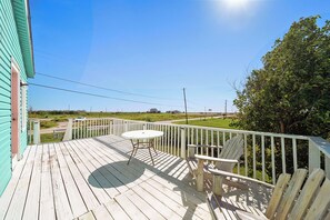 A wooden deck with a table and chairs, overlooking a grassy area with a road and distant buildings. - Colva Tides by Ryson Vacation Rentals (Galveston)