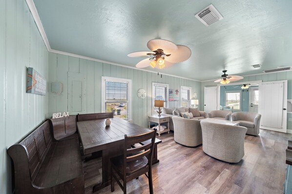 A dining area with a wooden table and benches, a sofa, and two ceiling fans. - Colva Tides by Ryson Vacation Rentals (Galveston)