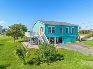 A turquoise house with a porch and a small deck, surrounded by green grass and trees. - Colva Tides by Ryson Vacation Rentals (Galveston)