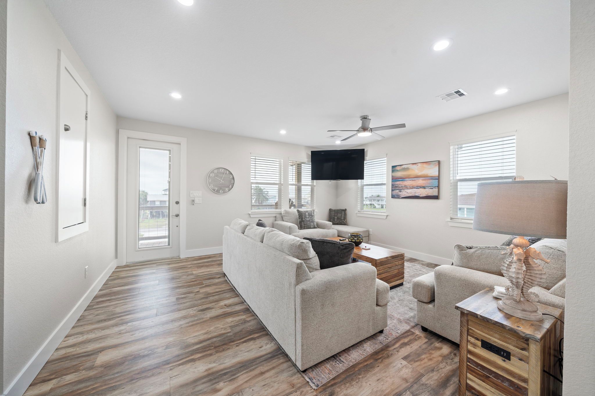 A spacious living room with a large sofa, a coffee table, and a television. There is a door leading to another room and a window with blinds.