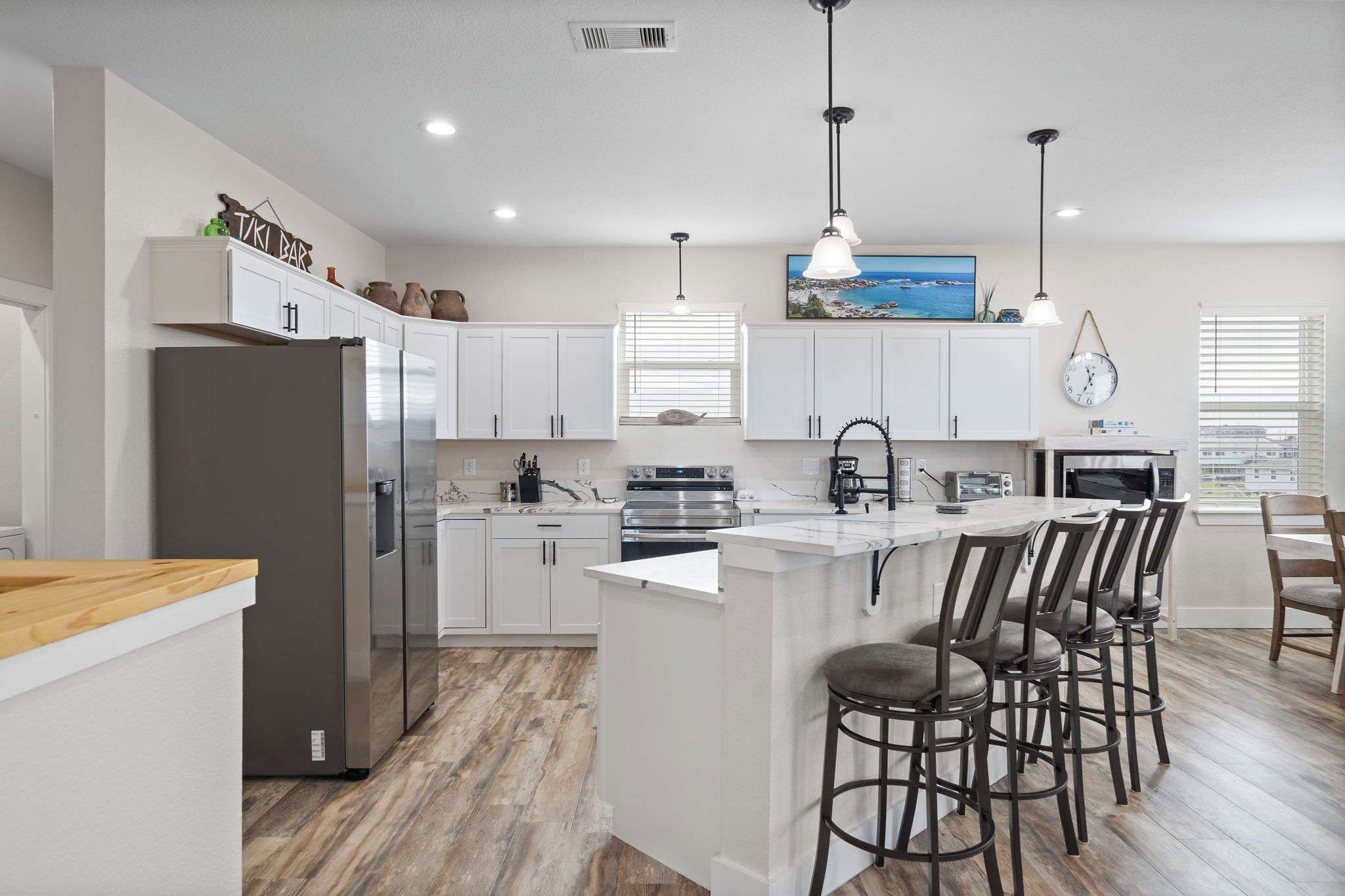 A modern kitchen with white cabinets, a stainless steel refrigerator, a central island with bar stools, and pendant lighting.