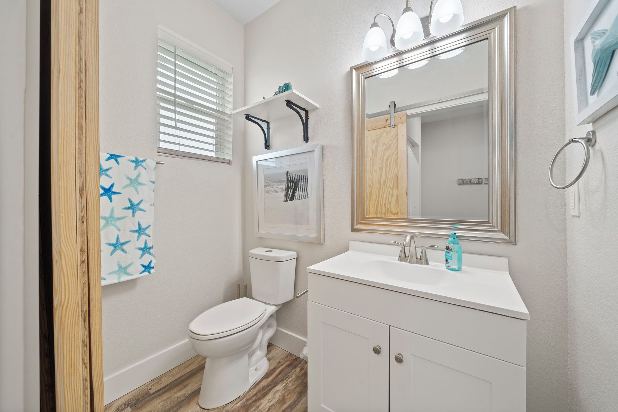 A bathroom with a toilet, sink, and mirror. There is a towel rack with a blue towel, a window with blinds, and a shelf with a picture frame.