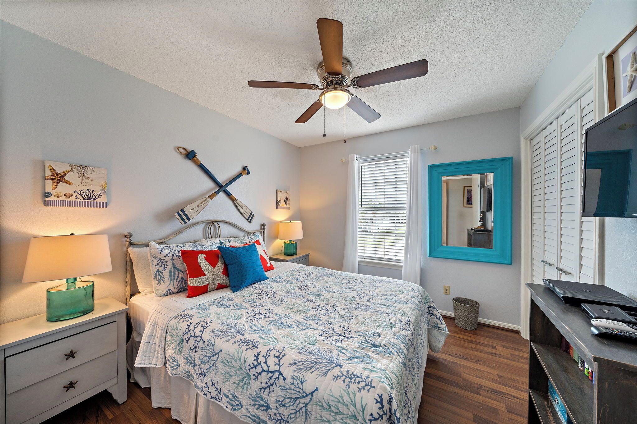A bedroom with a bed, nightstands, a ceiling fan, a window with blinds, a mirror, and a TV stand.