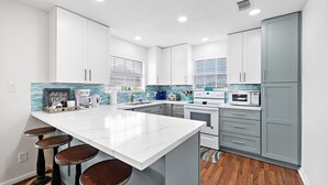 A modern kitchen with white and gray cabinetry, a large island with stools, and a tiled backsplash. - Island Breeze by Ryson Vacation Rentals (Galveston)