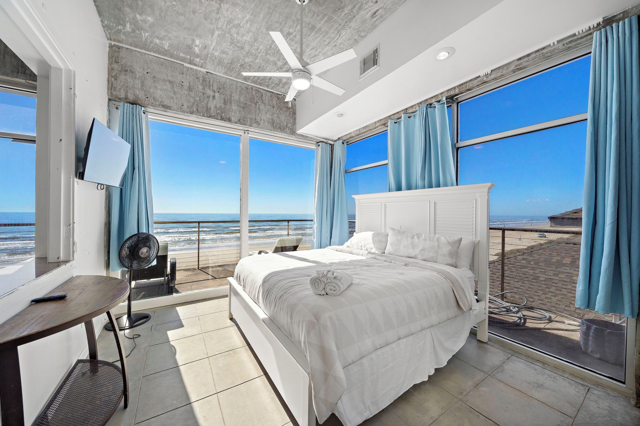 A bedroom with a large bed, a ceiling fan, a TV, and a view of the beach.