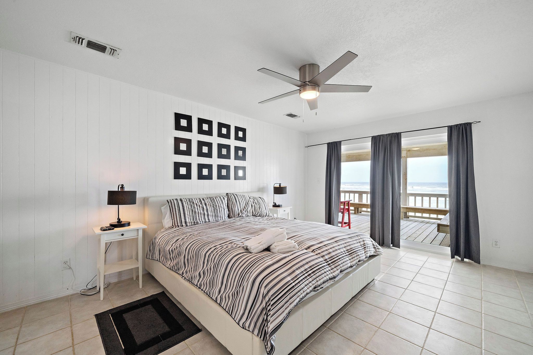 A bedroom with a bed, a ceiling fan, a nightstand, a wall-mounted picture frame, and a view of the beach through the window.