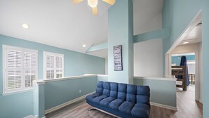 A room with a blue sofa, white shutters, a ceiling fan, and a wall-mounted sign listing city names. - The Krusty Krab by Ryson Vacation Rentals (Galveston)