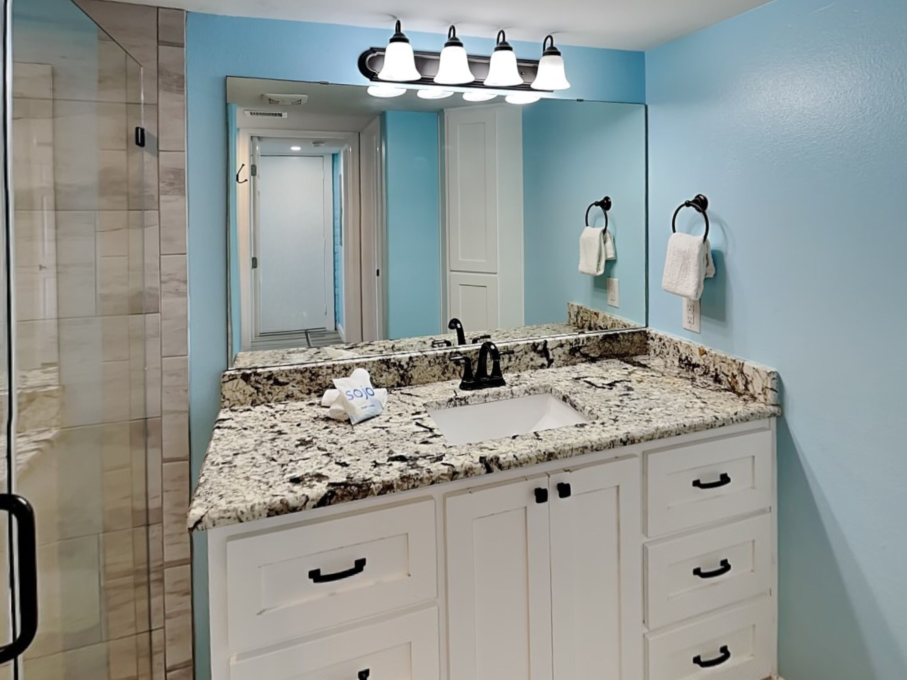 A bathroom with a granite countertop, white cabinets, a sink, and a mirror with lights above.