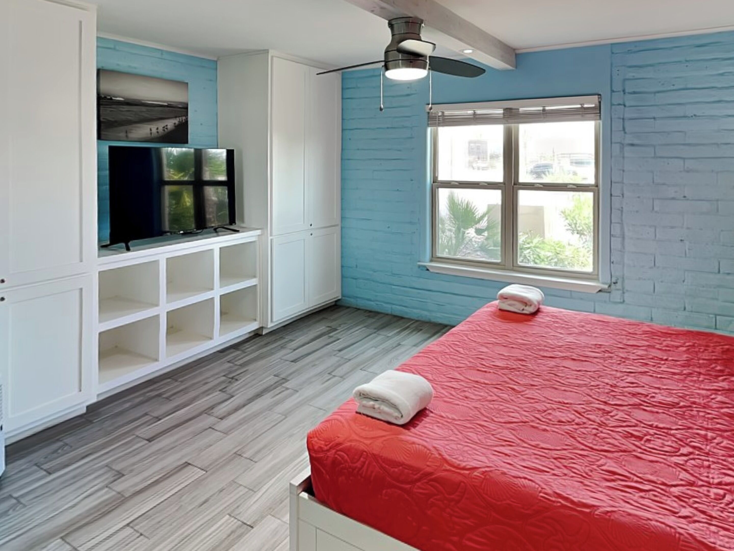 A bedroom with a red bedspread, a white wardrobe, a television on a shelf, and a window with a view of greenery.