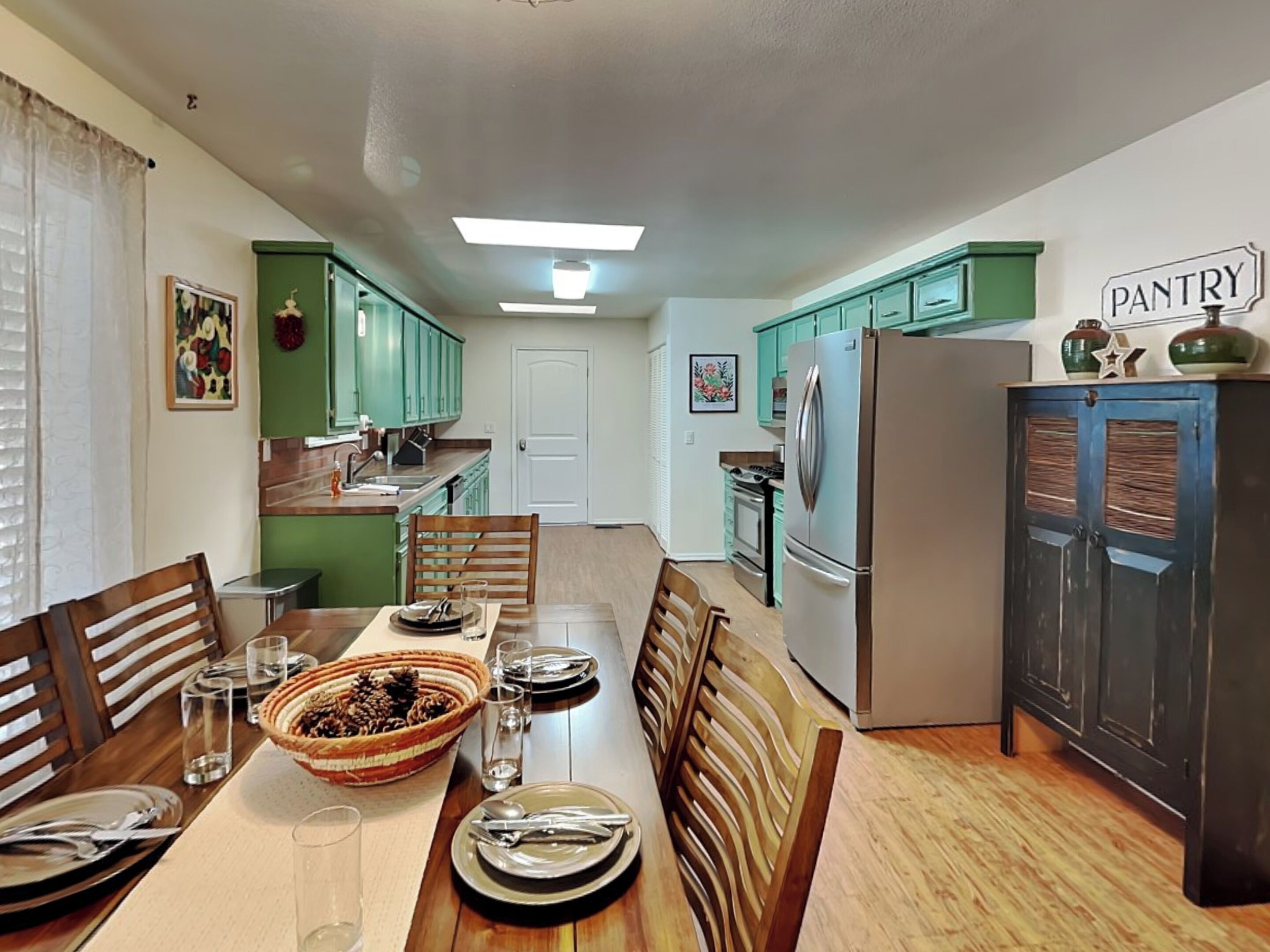 A dining area with a wooden table set for four, a kitchen with teal cabinets, and a pantry sign on the wall.